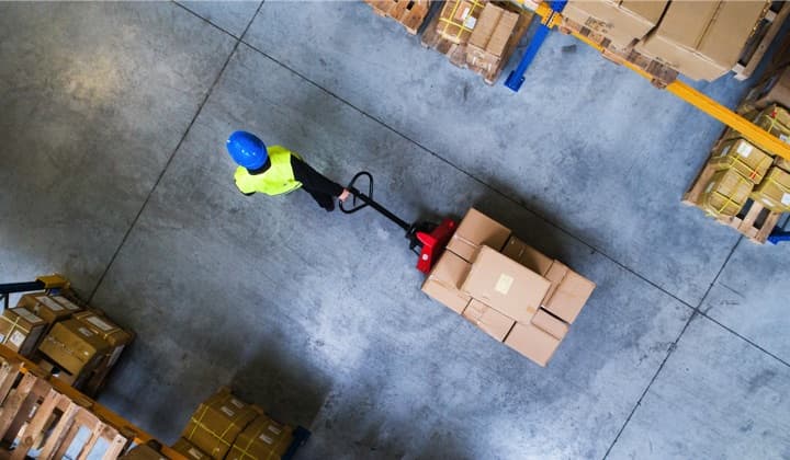 A birdeye view of a warehouse floor with a worker pulling a stack of boxes on a cart.