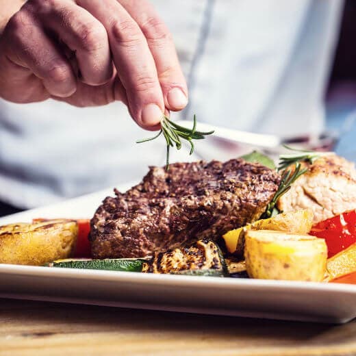 A photo of a chef’s hand adding finishing touches to a dish of steak with grilled vegetables.